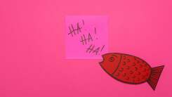 A cartoonish orange paper fish is affixed with a sticky note printed with "Ha! Ha! Ha!" on a pink background