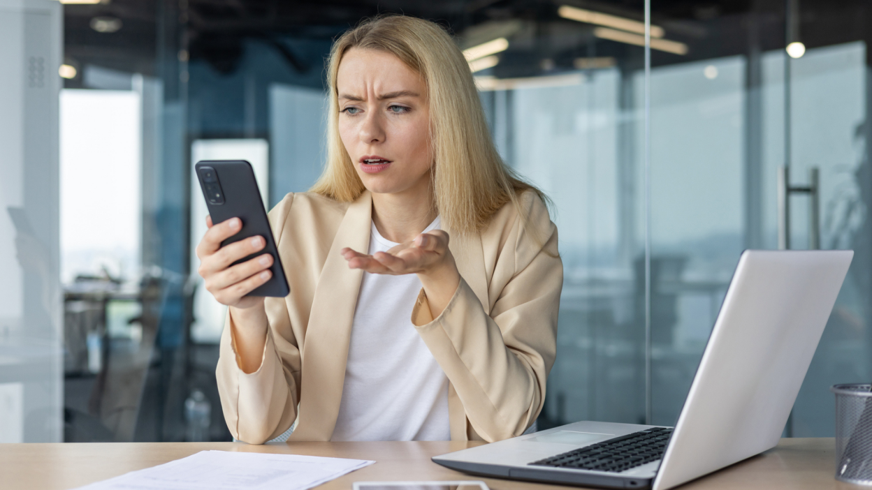 Worried young business woman working in the office at the laptop, holding the phone in her hands, anxiously looking at the screen - stock photo